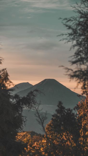 Stunning Sunrise View Of A Misty Volcano In Indonesia, Framed By Lush Trees. wallpaper preview
