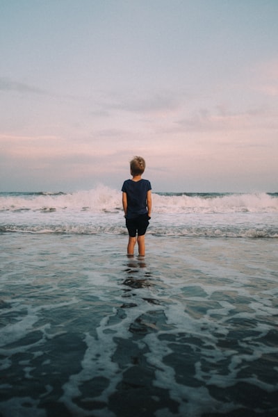 A Young Boy Stands In The Ocean Waves At Sunset. wallpaper preview
