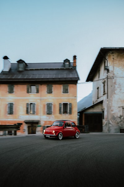 A Red Vintage Car Drives Past Old Buildings. wallpaper preview