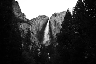 Yosemite Falls Thunders Down A Sheer Granite Face, Framed By Dark Conifers In This High-contrast Black And White. The Bright Plume Of Water Cuts Through The Shadowed Valley, Where The Upper Fall Plunges Into Mist Before Reaching The Lower Cascade. A Study In Light And Stone, Captured In Classic Monochrome. wallpaper preview