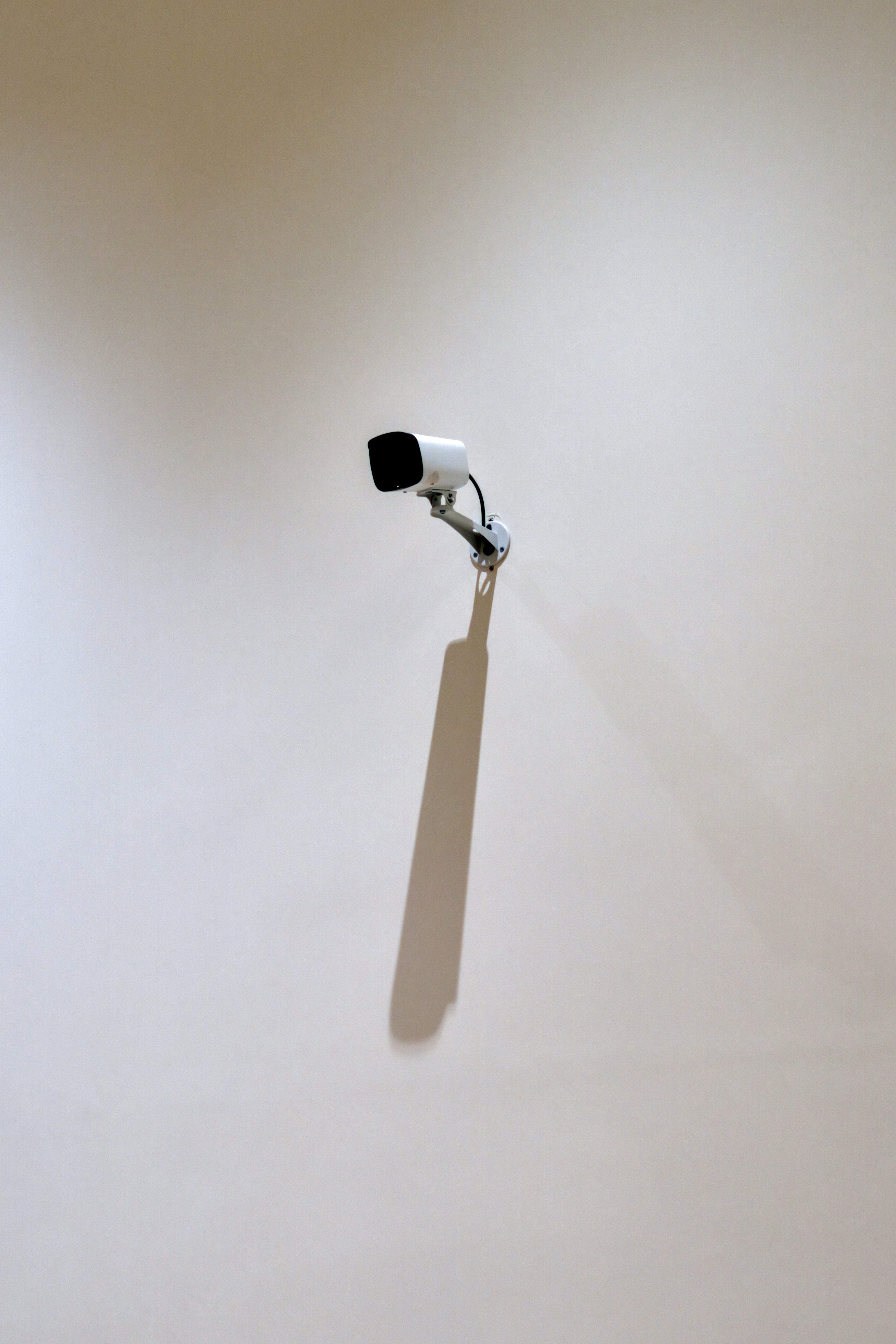A Security Camera Mounted On A Blank Wall Casts A Long Shadow Under Soft Indoor Light. Surrounded By Empty Space, The Ordinary Object Becomes A Minimalist Study Of Surveillance, Geometry, And Presence. HD mobile wallpaper