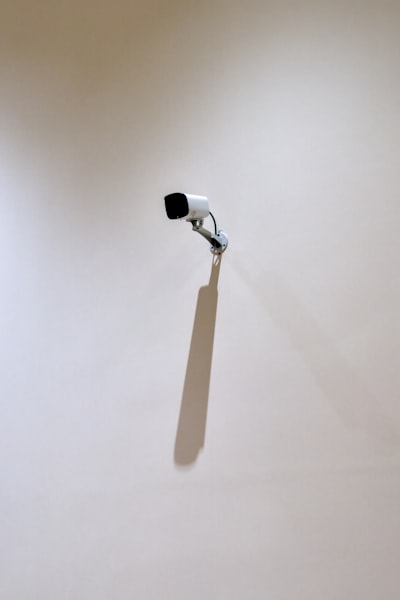 A Security Camera Mounted On A Blank Wall Casts A Long Shadow Under Soft Indoor Light. Surrounded By Empty Space, The Ordinary Object Becomes A Minimalist Study Of Surveillance, Geometry, And Presence. wallpaper preview