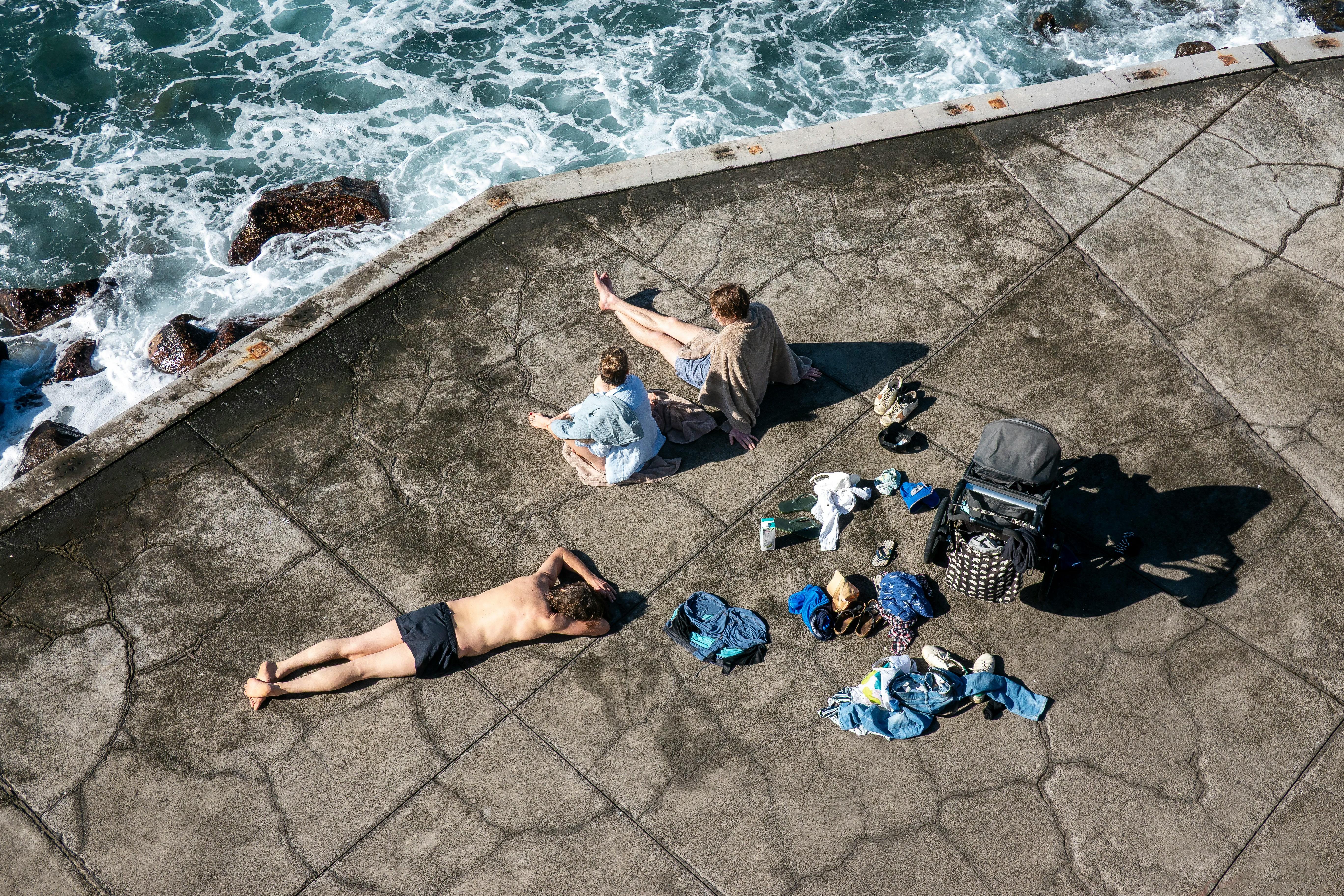 An Aerial View Of People Relaxing By The Ocean Near Funchal, Portugal. A Man Lies On The Ground, While Two Women Sit Together. Various Items Of Clothing And A Stroller Are Scattered Around Them. HD mobile wallpaper
