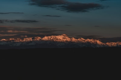 A Dramatic Alpine Mountain Range Called "Alpstein Region" Rises Across The Horizon, Its Snow-covered Peak "Säntis" Is Illuminated By The Warm Golden Light Of Dawn. Dark Foreground Silhouettes Contrast With The Glowing Ridges, While A Deep Blue Sky Filled With Scattered Clouds Creates A Moody And Atmospheric Backdrop. wallpaper preview