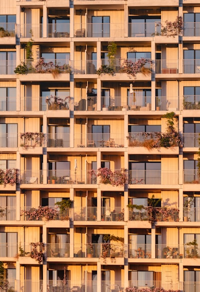 Close-up Facade Of A Modern Residential Apartment Building Glowing In Warm Golden Sunset Light, Showcasing A Repeating Grid Of Private Balconies Filled With Lush Plants, Flowers, And Greenery. wallpaper preview