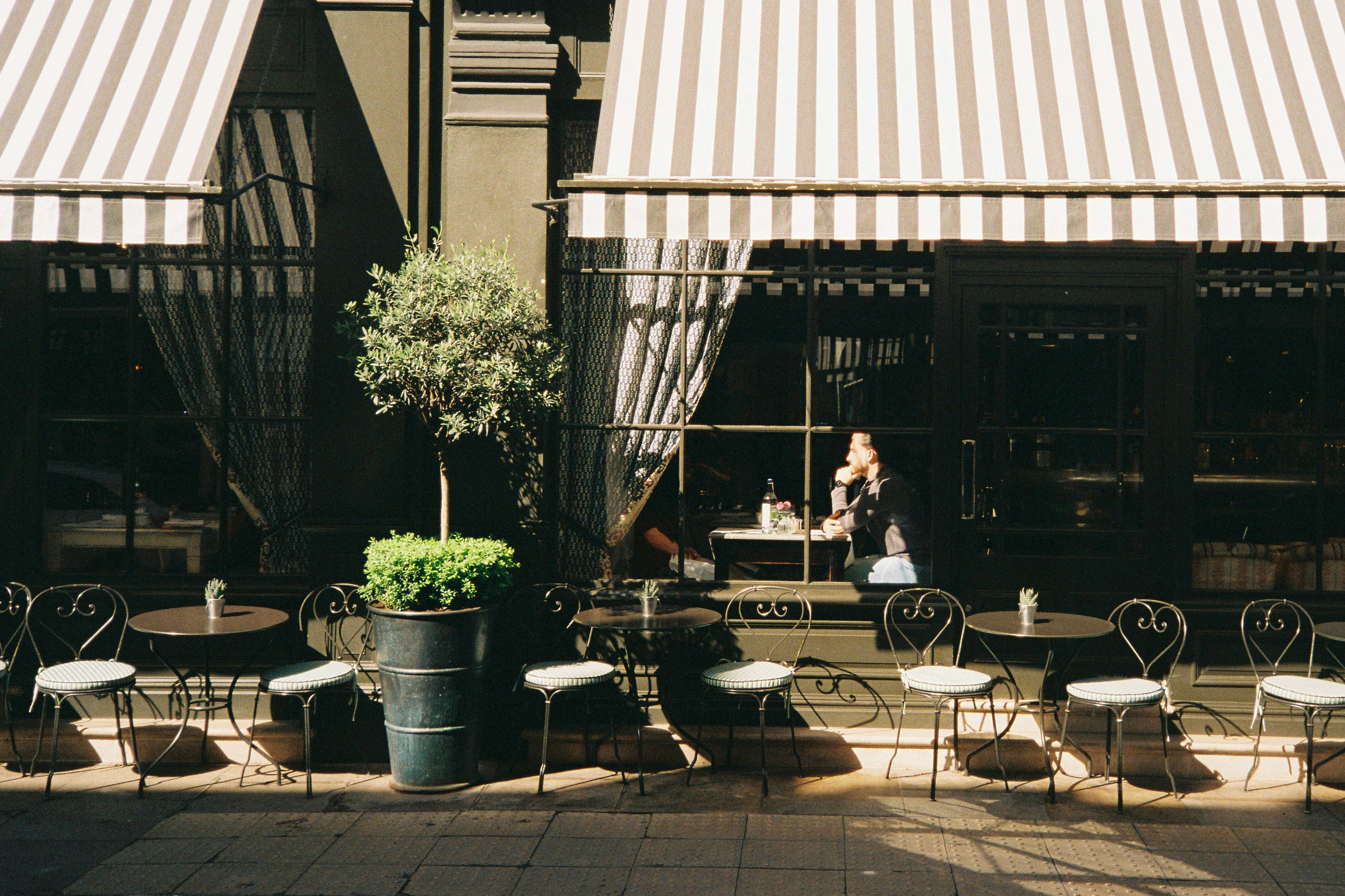 A Street View Of A London Restaurant Café. There Is A Row Of Small Empty Tables Outside And A Striped Awning. A Man Is At The Window Sitting At A Table With His Hand Under His Chin. There Is A Shaft Of Sunlight Across Him. Shot On Film. HD mobile wallpaper