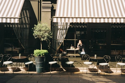 A Street View Of A London Restaurant Café. There Is A Row Of Small Empty Tables Outside And A Striped Awning. A Man Is At The Window Sitting At A Table With His Hand Under His Chin. There Is A Shaft Of Sunlight Across Him. Shot On Film. wallpaper preview