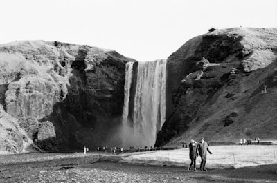 People Walk Near A Large Waterfall In A Mountainous Landscape. wallpaper preview
