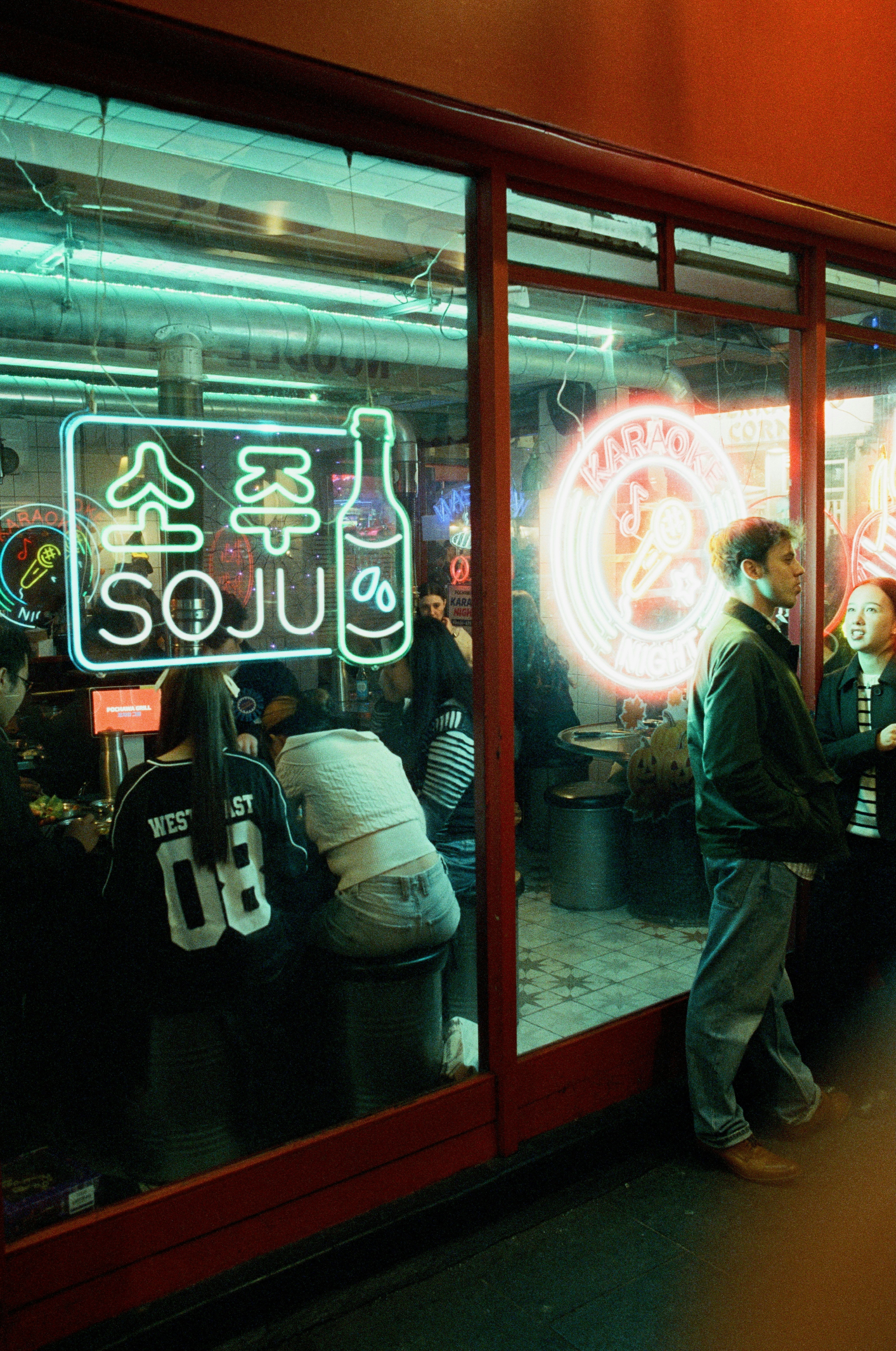 Neon Soju And Karaoke Signs Glow In A Window As People Gather Outside A Bar At Night. HD mobile wallpaper