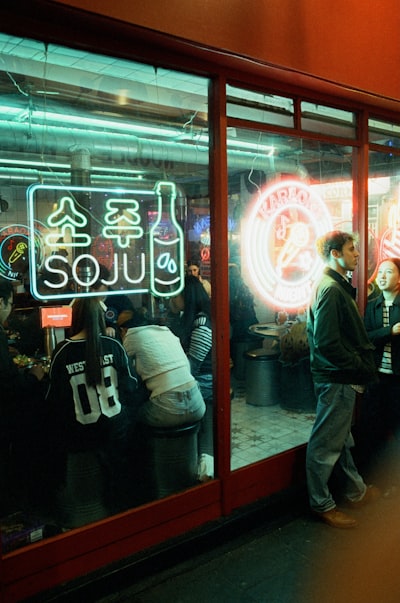 Neon Soju And Karaoke Signs Glow In A Window As People Gather Outside A Bar At Night. wallpaper preview