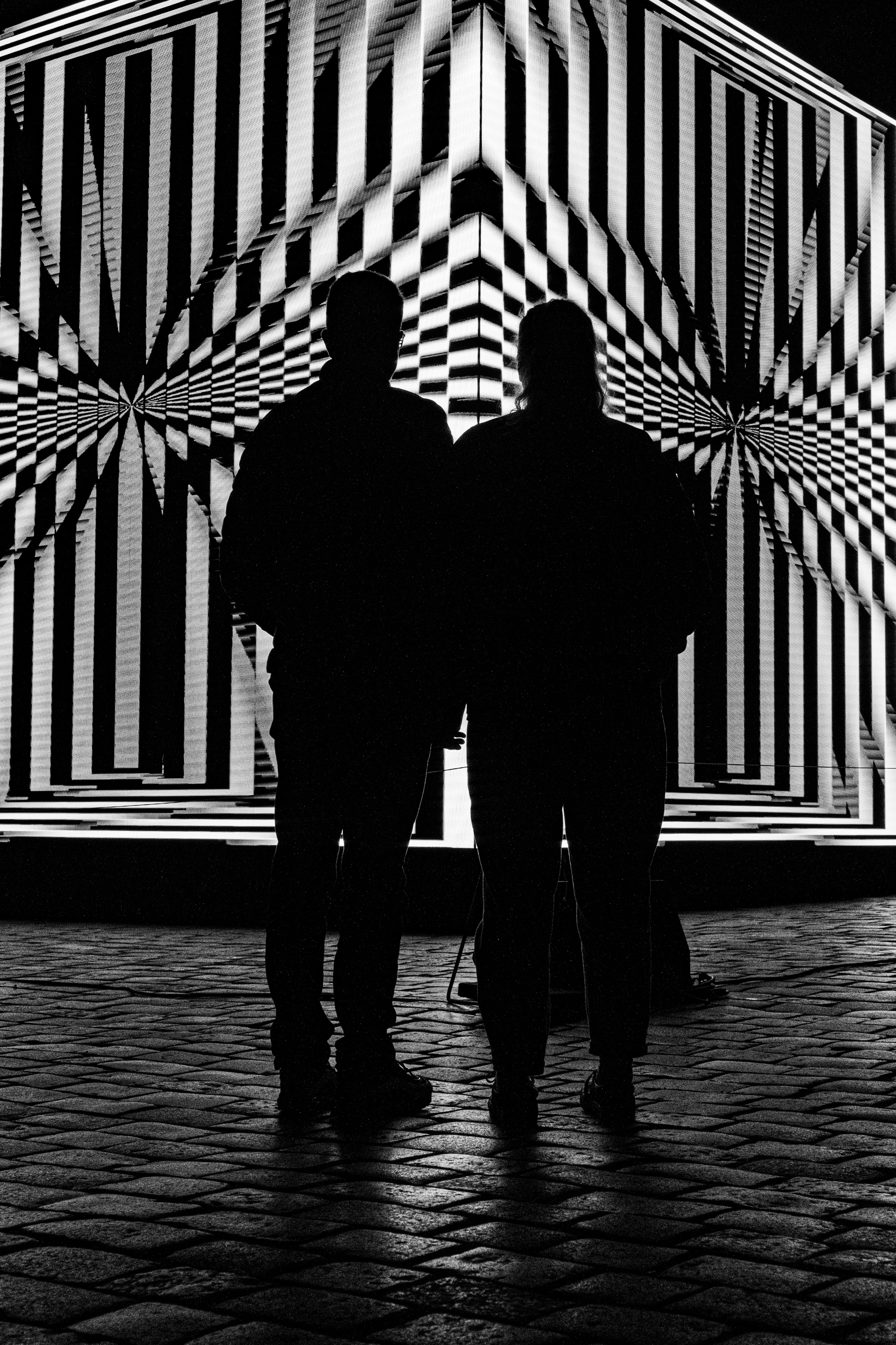 Two People Stand In Silhouette In Front Of A High-contrast Geometric Light Installation. The Repeating Black And White Pattern Creates A Strong Optical Illusion And A Dramatic Urban Night Mood. HD mobile wallpaper