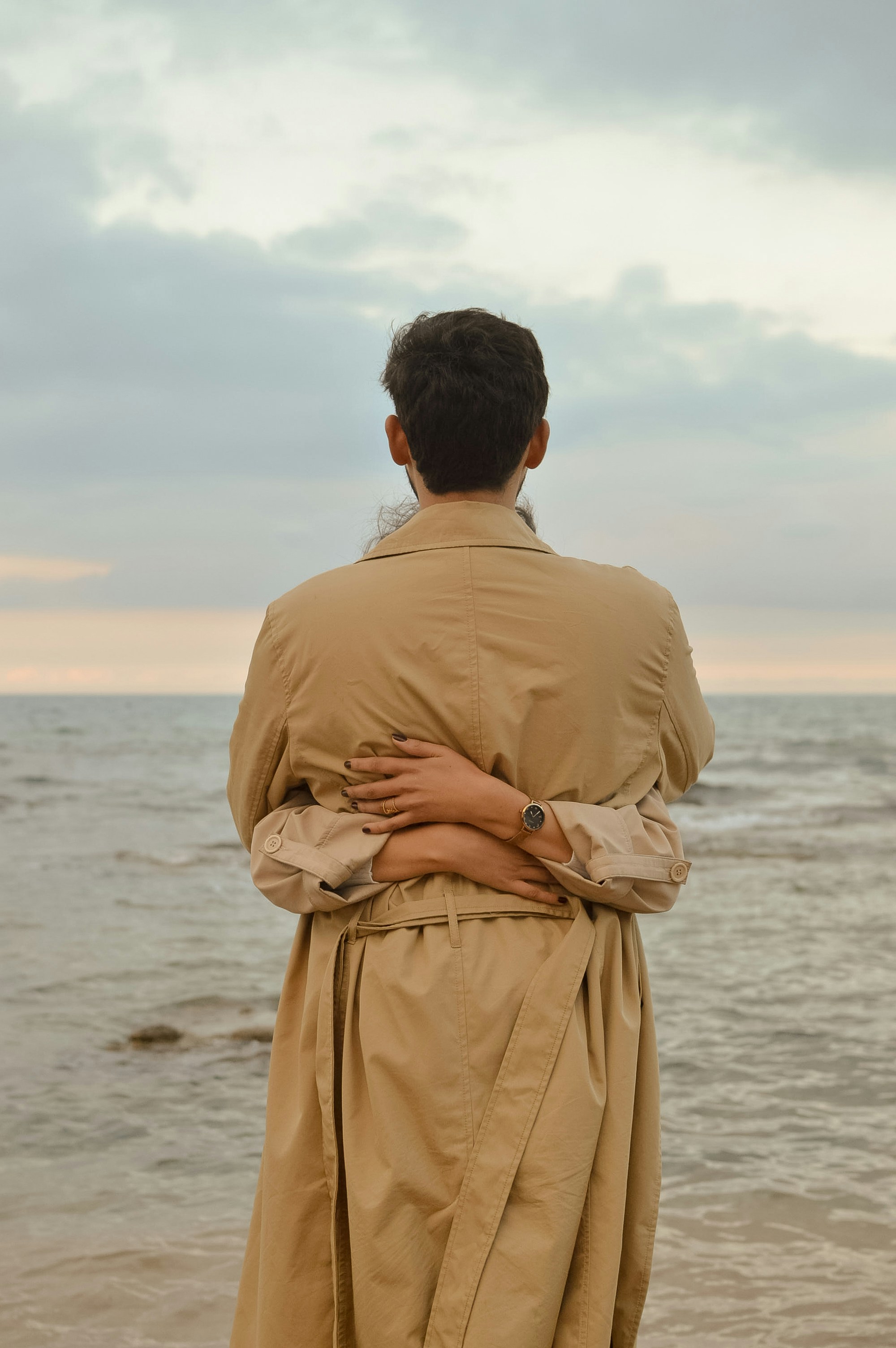 Person In Trench Coat Embraced By Another By The Ocean. HD mobile wallpaper