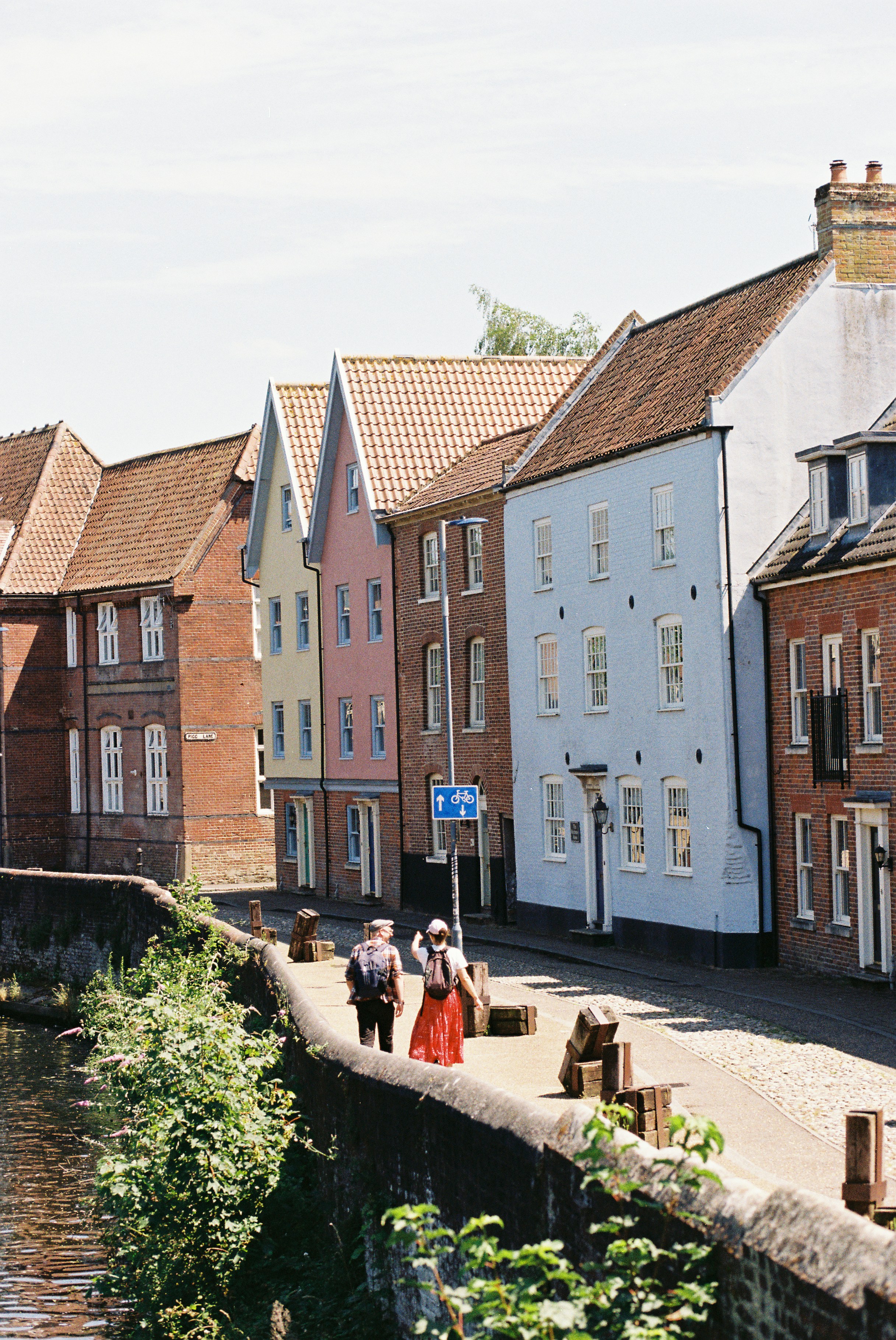 Colourful Gabled Cottages Line A Cobbled Riverside Lane Where Two Walkers Pass In Summer Light. HD mobile wallpaper