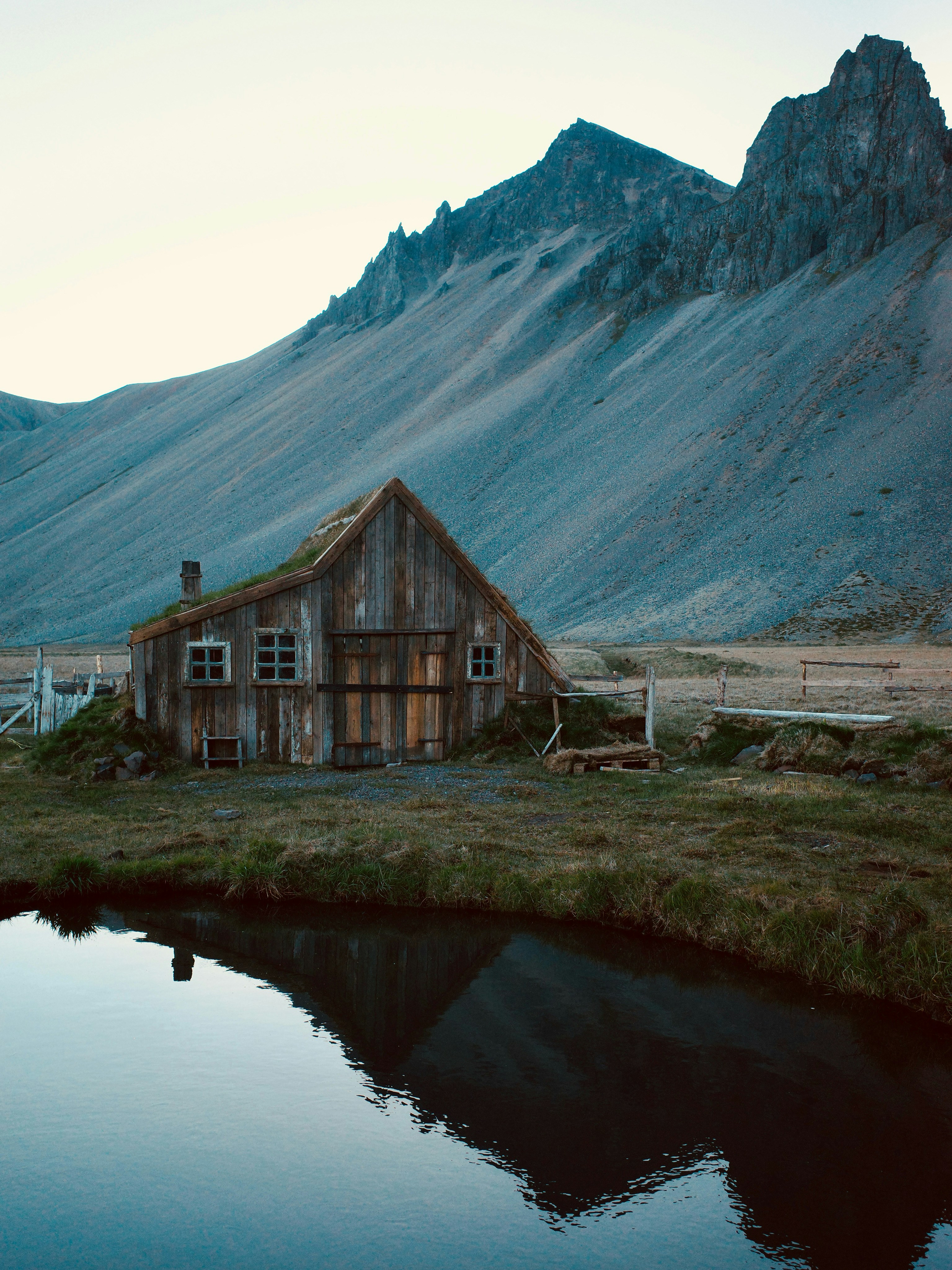 A Rustic Wooden House Reflects In Water With Mountains Behind. HD mobile wallpaper