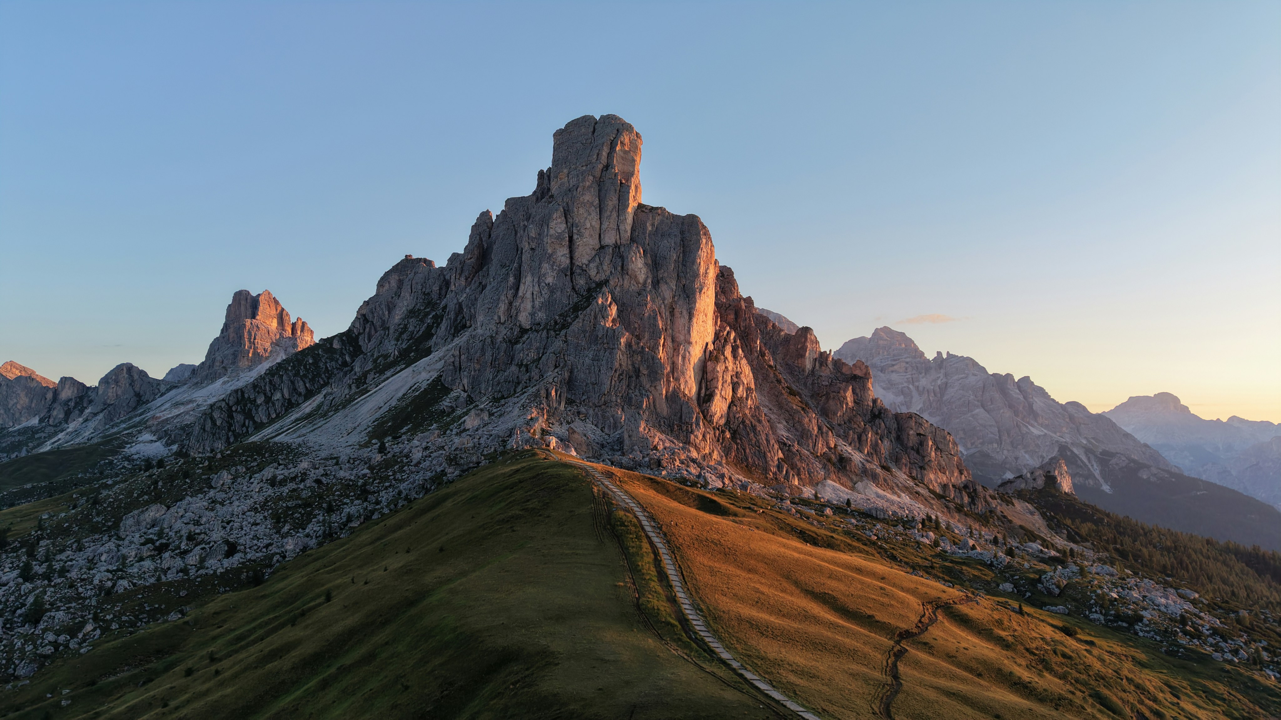 A Narrow Hiking Trail Winding Up A Grassy Slope Toward Jagged Rocky Peaks. HD mobile wallpaper
