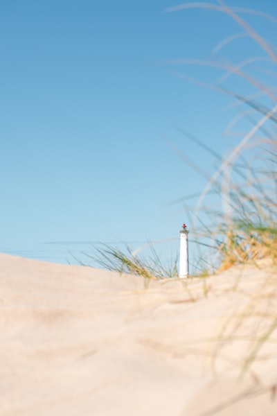 A White Lighthouse Behind Sand Dunes With Tall Grass Under A Clear Sky wallpaper preview