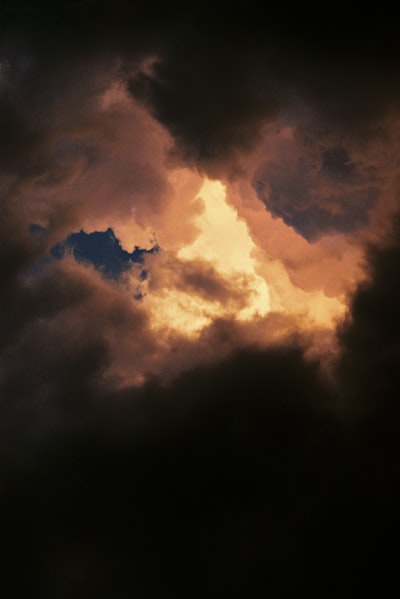 Grainy 35mm Frame Of Heavy Cloud Filling The Whole Image, Almost Entirely Dark Except For A Jagged Tear At Centre Where The Setting Sun Blazes Through In White-hot Gold. Rusty Brown And Violet Tones Bloom Around The Opening; The Lower Third Falls Away Into Near Black. Soft Focus, Visible Grain, Painterly And Dramatic. wallpaper preview