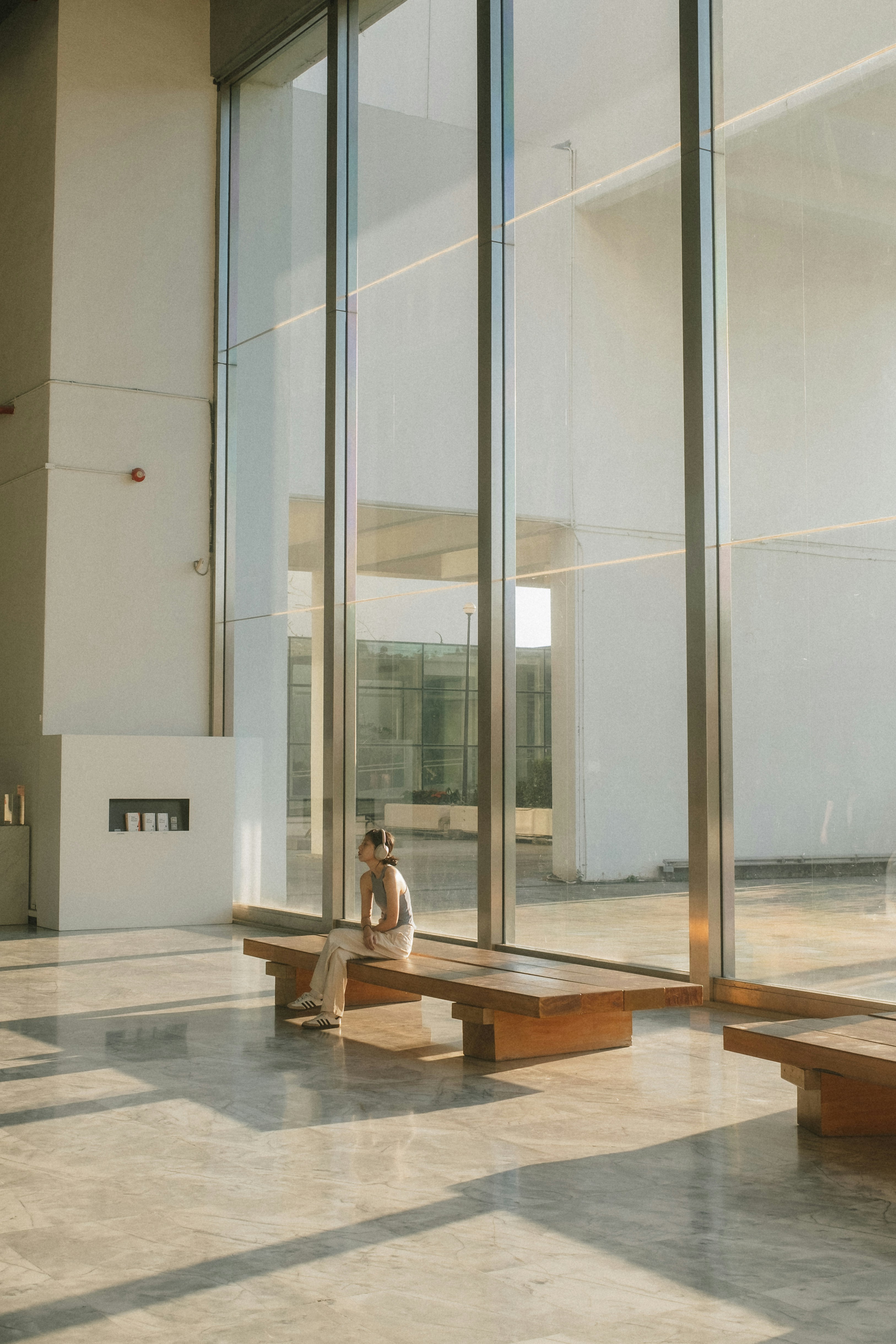 A Person Wearing Headphones On A Wooden Bench In A Sunlit Modern Lobby HD mobile wallpaper