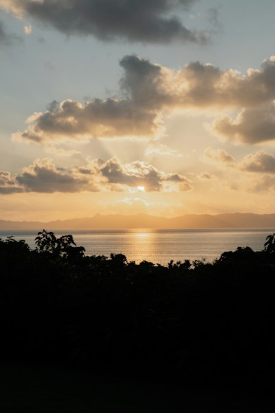 Low Sun Glowing Behind Clouds Over A Calm Sea And Distant Mountain Silhouettes wallpaper preview