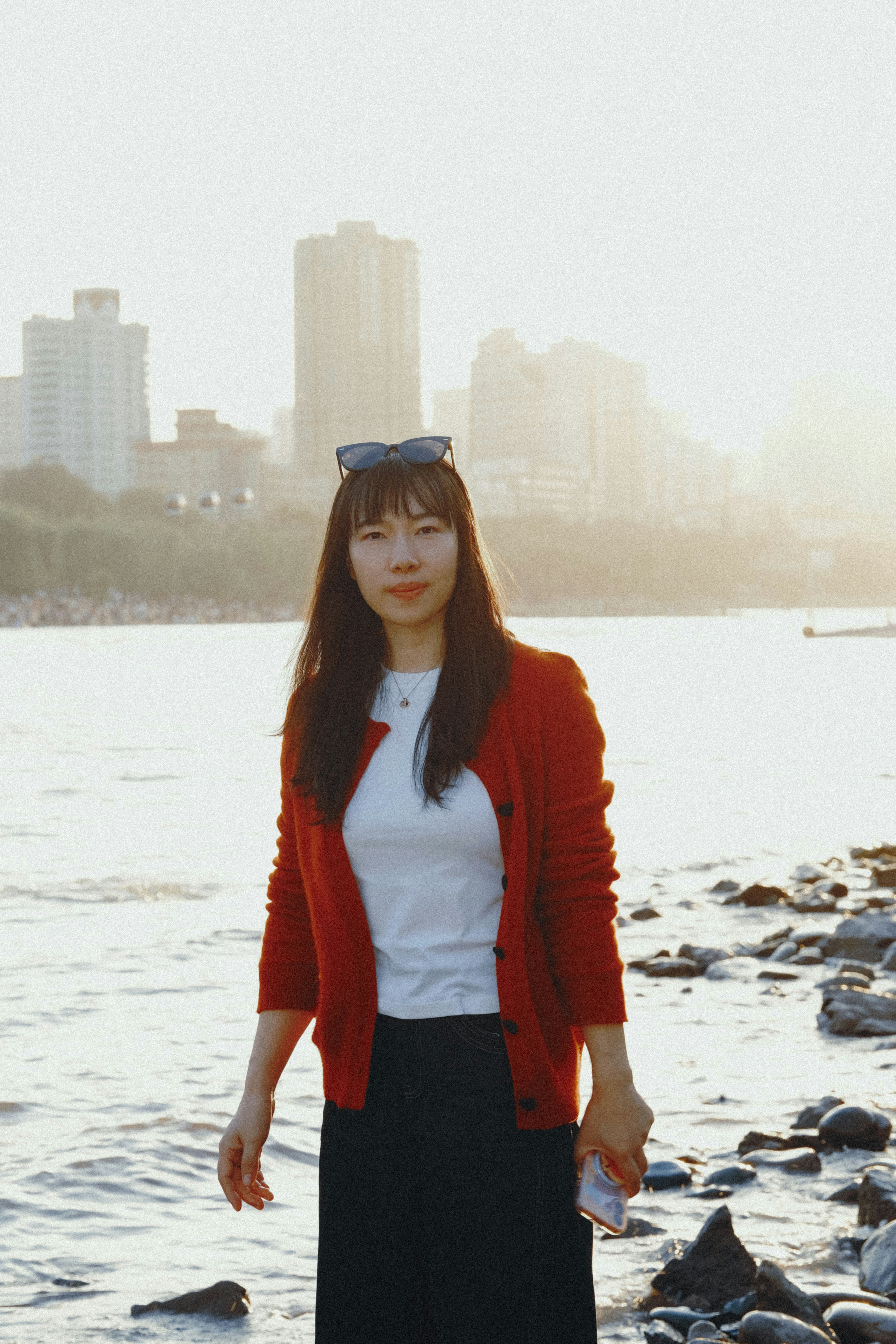 A Woman In A Red Cardigan Standing By A River With City Buildings In The Background HD mobile wallpaper