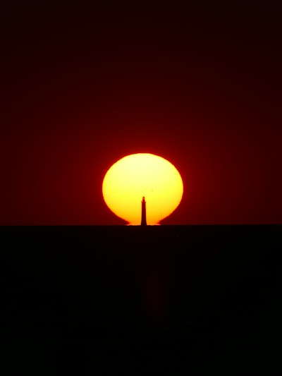 A Lighthouse Silhouette Centered Against A Large Low Sun On The Horizon wallpaper preview