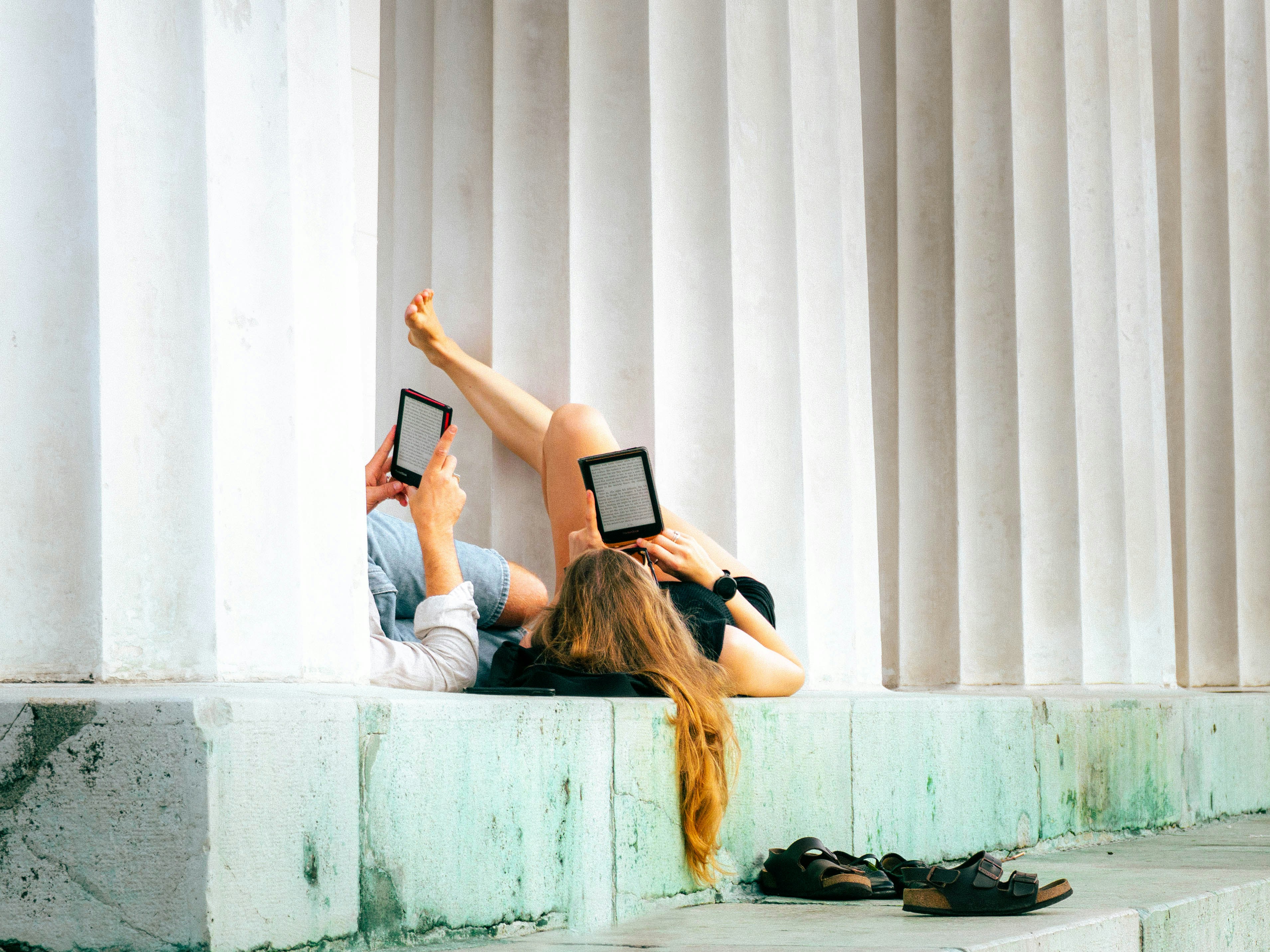 Two People Reading E-readers While Reclining Against White Fluted Columns On A Stone Ledge HD mobile wallpaper