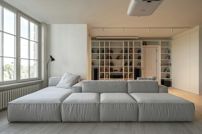Daylight Fills A Minimalist Living Room With A Large Gray Modular Sofa And A Wall-to-wall White Bookcase. The Custom Library And Double-sided Seating Organize An Open-plan Apartment Overlooking Kyiv. Interior Design By PAINTIT; Photography By Andriy Shurpenkov. wallpaper preview