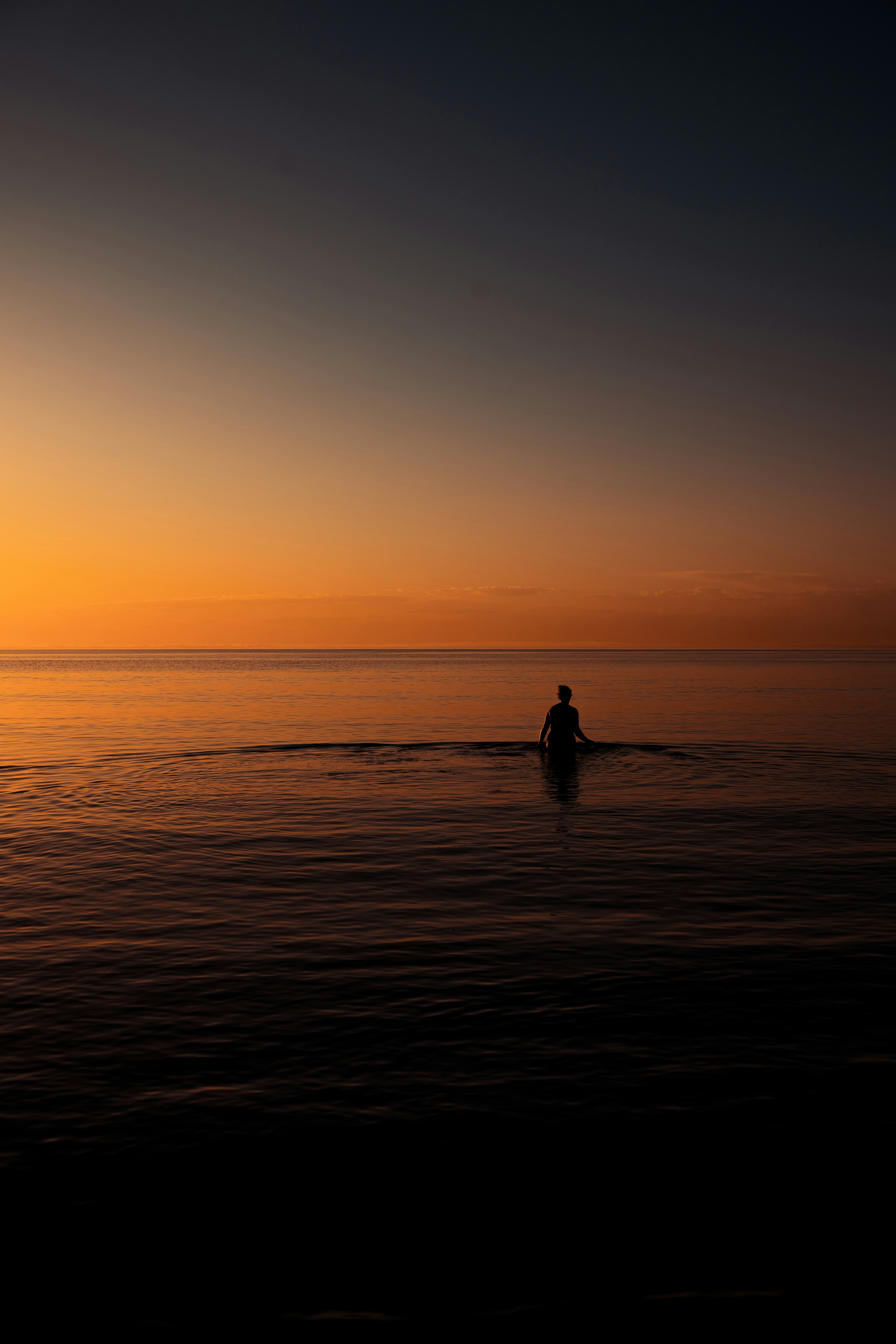 A Person In Silhouette Wading In Calm Water Under A Warm Orange Sky HD mobile wallpaper