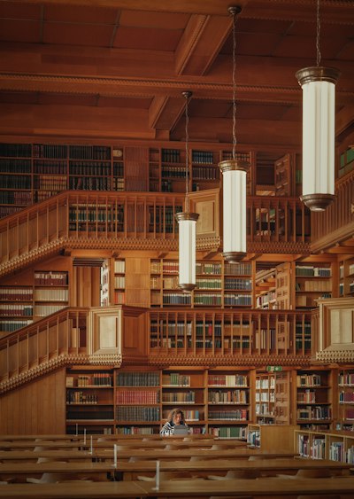 A Person Working On A Laptop In A Large Wooden Library With Tiered Bookshelves wallpaper preview
